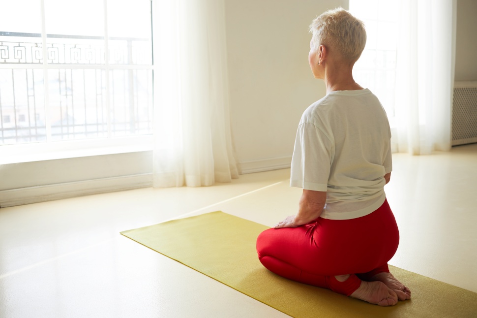 Yoga, meditation, zen and peace concept. Back view of short haired middle aged woman sitting barefooted on mat in front of large window, meditating, watching sunrise in calm peaceful atmosphere
